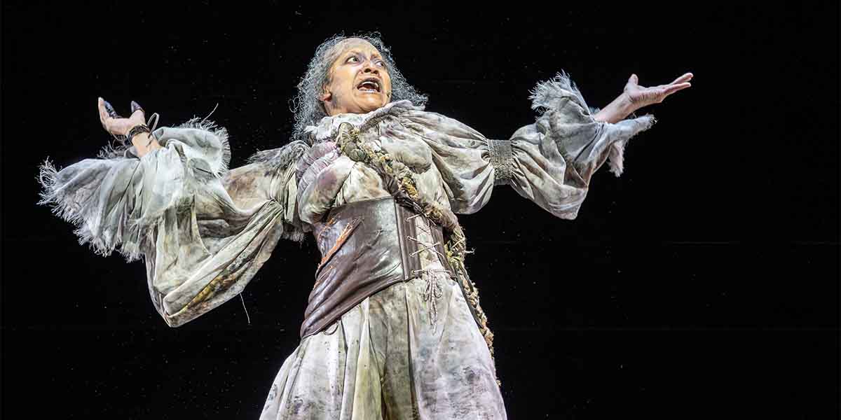 Melanie La Barrie performs passionately in a tattered costume, arms outstretched against a dark background.