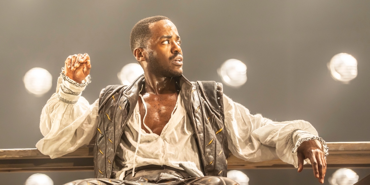 Ncuti Gatwa, smiling, poses with hands on hips, wearing a patterned shirt against a blurred background.