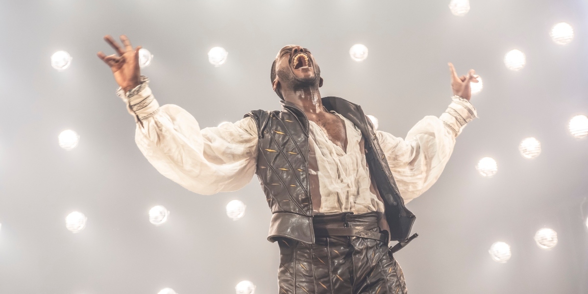Ncuti Gatwa poses confidently, wearing a blue suit with a white shirt, against a blurred background.