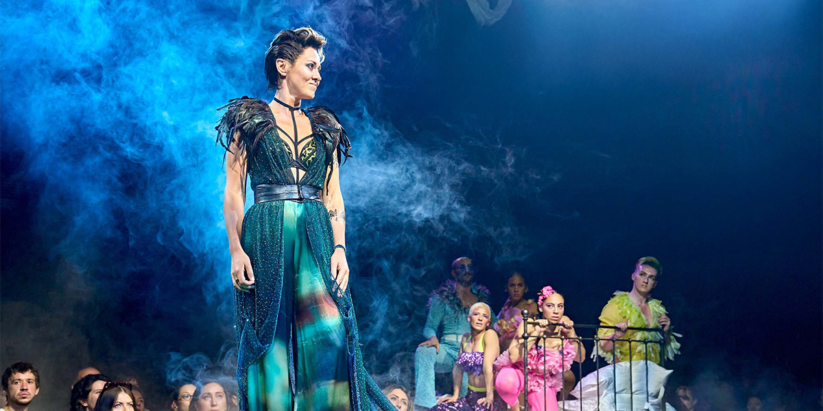 A performer on stage in an elaborate costume with a dramatic blue and green design, surrounded by a blue mist, with company in the background.