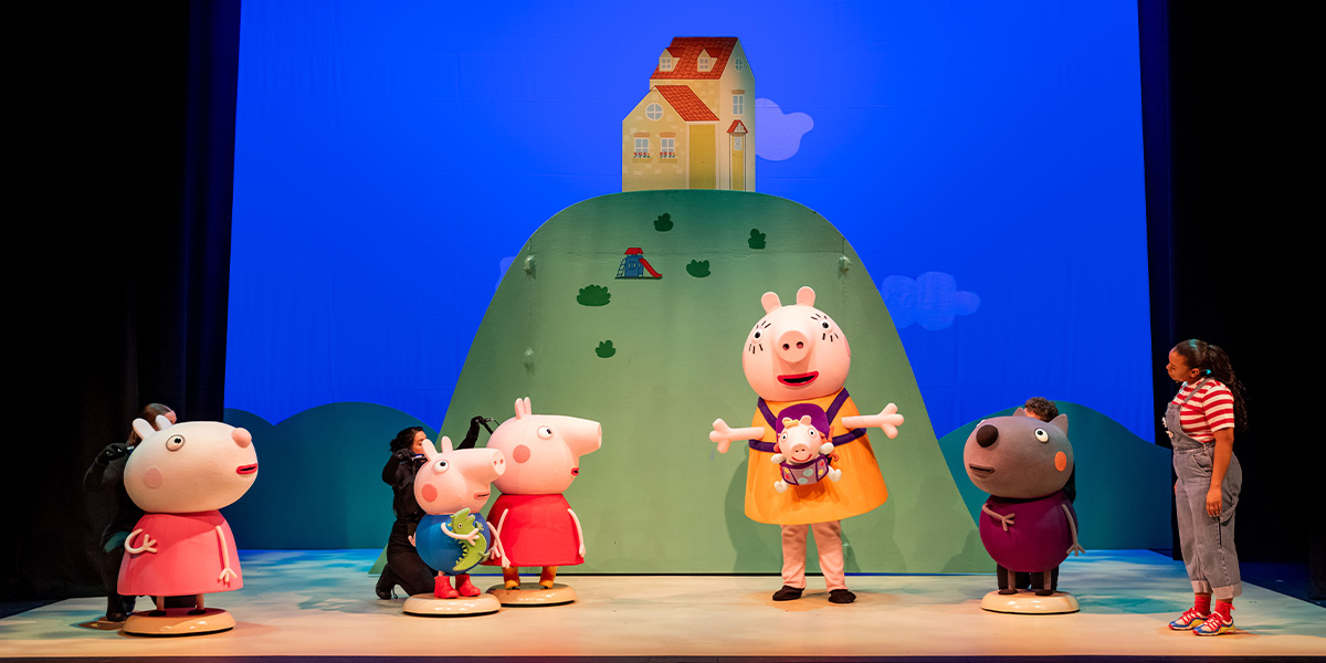 Peppa Pig and friends perform on stage, with a colourful backdrop of a hill and house.