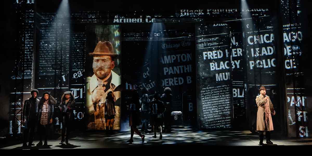 A group of performers stands on stage, with projected newspaper headlines and a spotlight on one speaker.