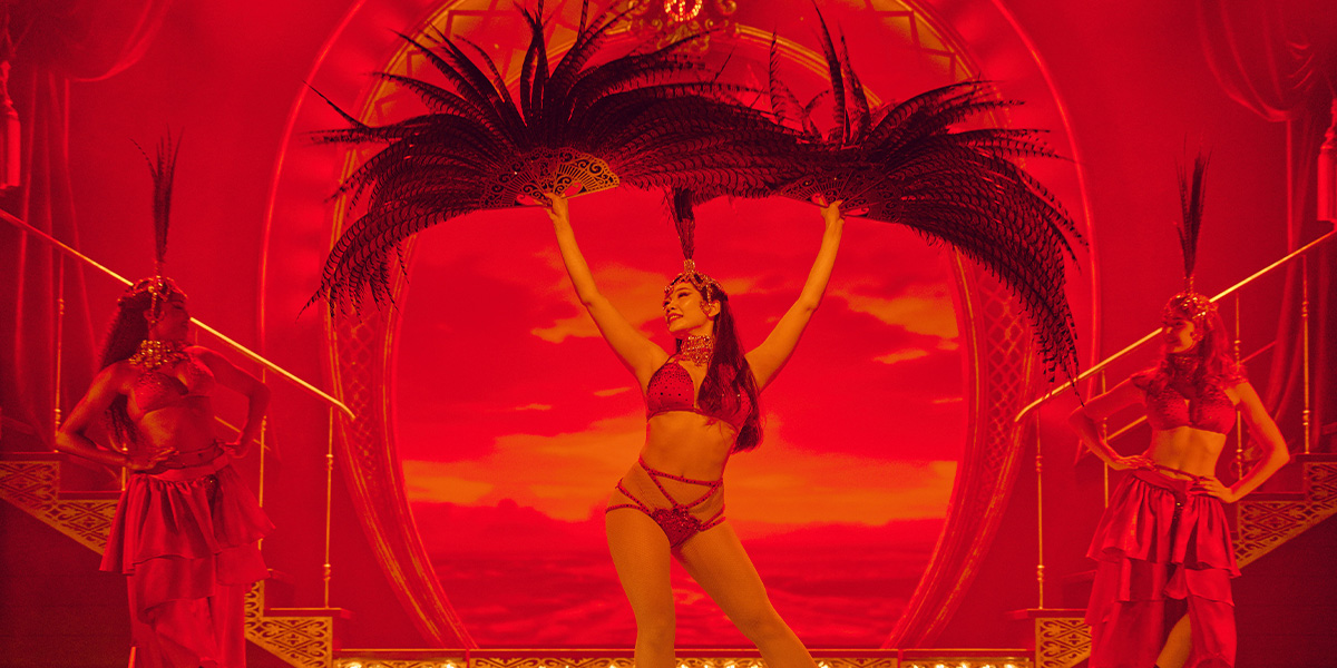 Performers in lavish costumes on a stage with dramatic red lighting, set against an ornate circular backdrop. The central figure holds a large feathered headdress aloft.