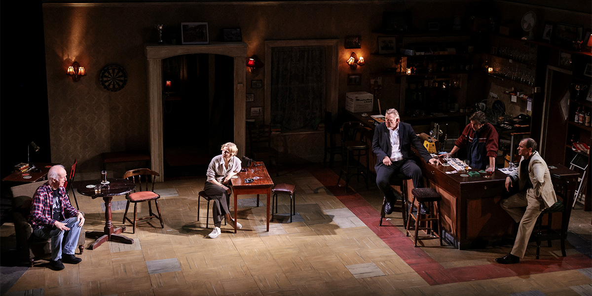 A group of five actors stands together, dressed in casual clothing, against a dimly lit rustic backdrop.