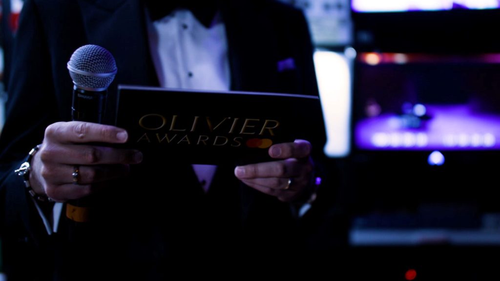 A person in a tuxedo holds an Olivier Awards card and microphone, with blurred background.