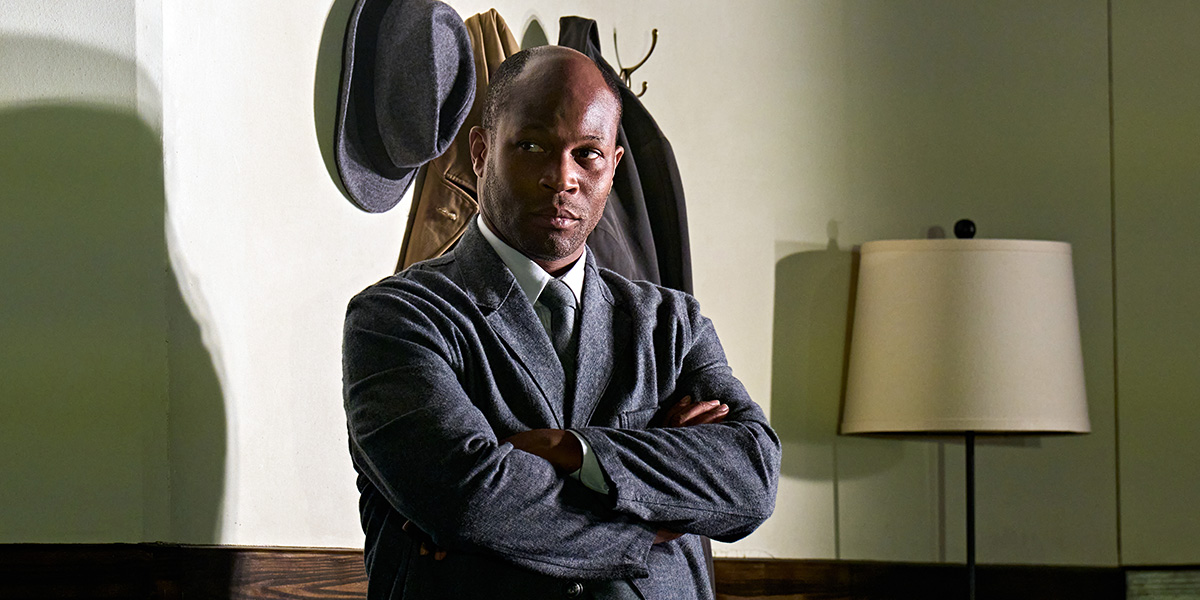 Person standing with arms crossed, wearing a formal suit, in a room with hats hanging on the wall and a lamp on a side table.