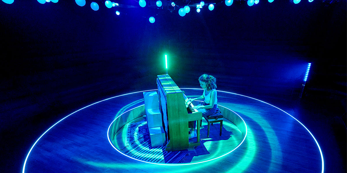 A person is playing a wooden piano surrounded by circular blue neon lights on a dark stage, illuminated also by a soft green light from above.