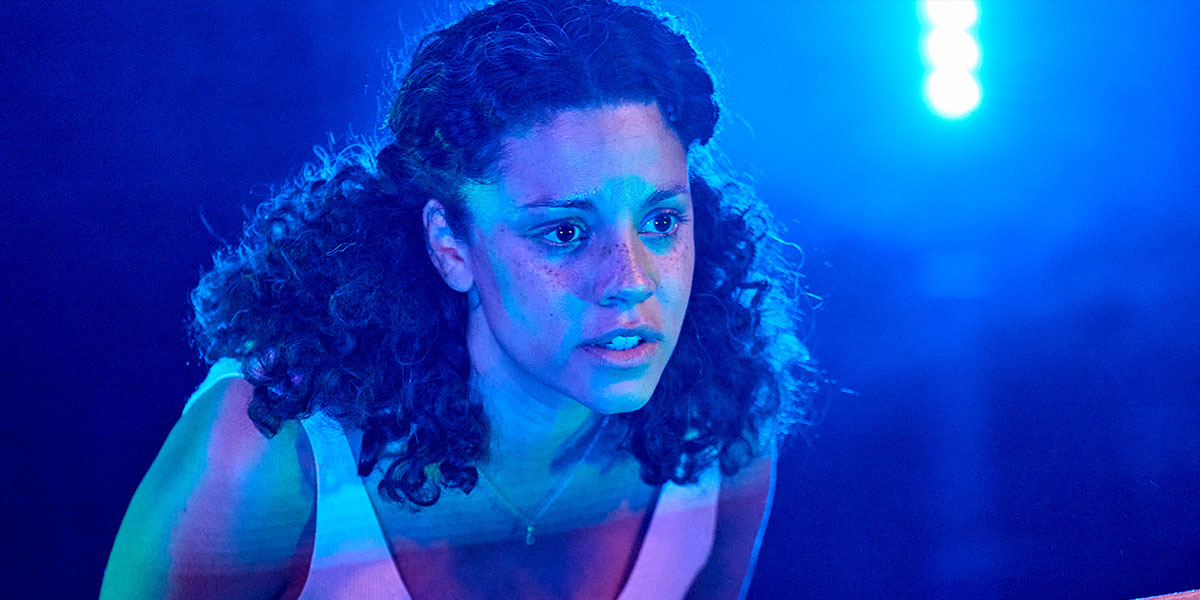 A person with curly brown hair is sitting on stage bathed in blue light.