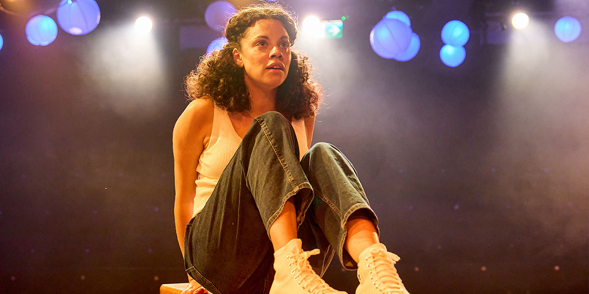 Person seated on stage under spotlight, looking contemplative, in a casual outfit with denim jeans and white sneakers.