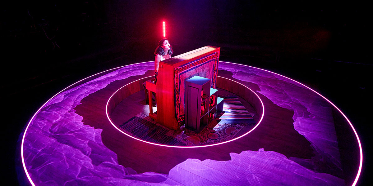 A performer at a piano on stage, surrounded by a circular purple light and a striking red beam from above.