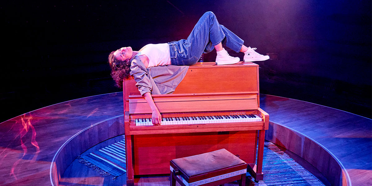 Person lounging on top of a grand piano on a stage with dramatic lighting.