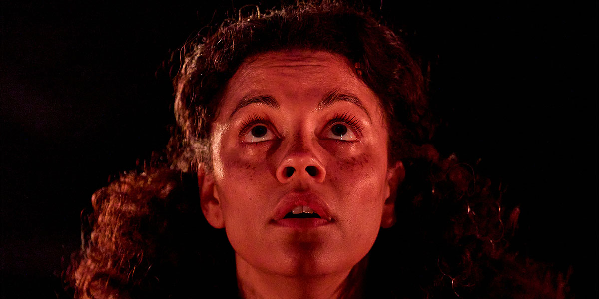 A person with curly hair gazes upward with an intense expression, illuminated by a warm, reddish light.