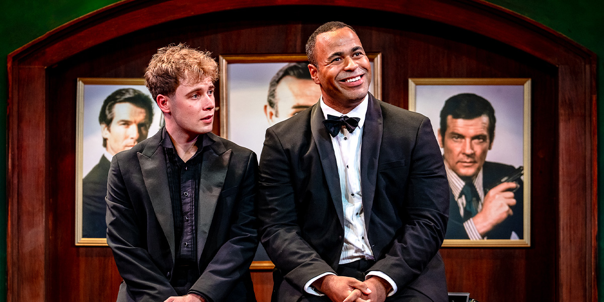 Two individuals are sitting side by side at a staged event, both dressed in formal attire. Behind them are paintings of famous James Bond actors displayed on the wall.