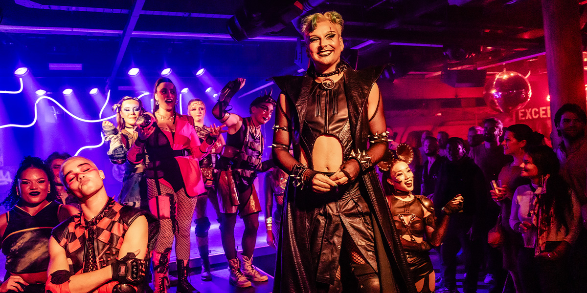 Jan Sport, dressed in period costume, poses centre stage with actors in similar attire, surrounded by theatrical decor.