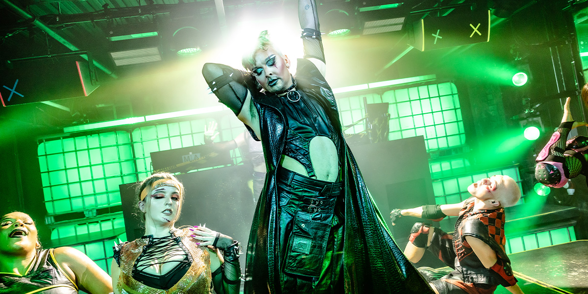 A man dressed as Oscar Wilde, wearing a dark suit and bow tie, poses thoughtfully against a textured backdrop.