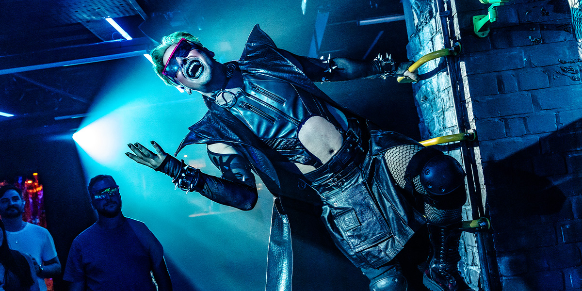 Jan Sport, dressed in a green velvet suit, poses dramatically with a quill in hand against a dark backdrop.