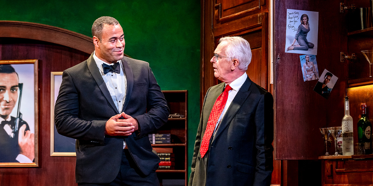 Two people conversing on a stage set to resemble a cosy office, with memorabilia and posters on the walls.