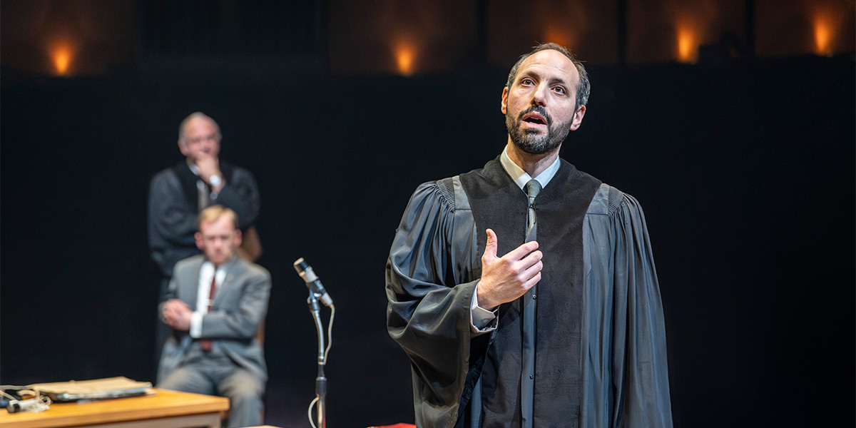 Philip Arditti, in a black robe, gestures while speaking, with two seated figures in the background.
