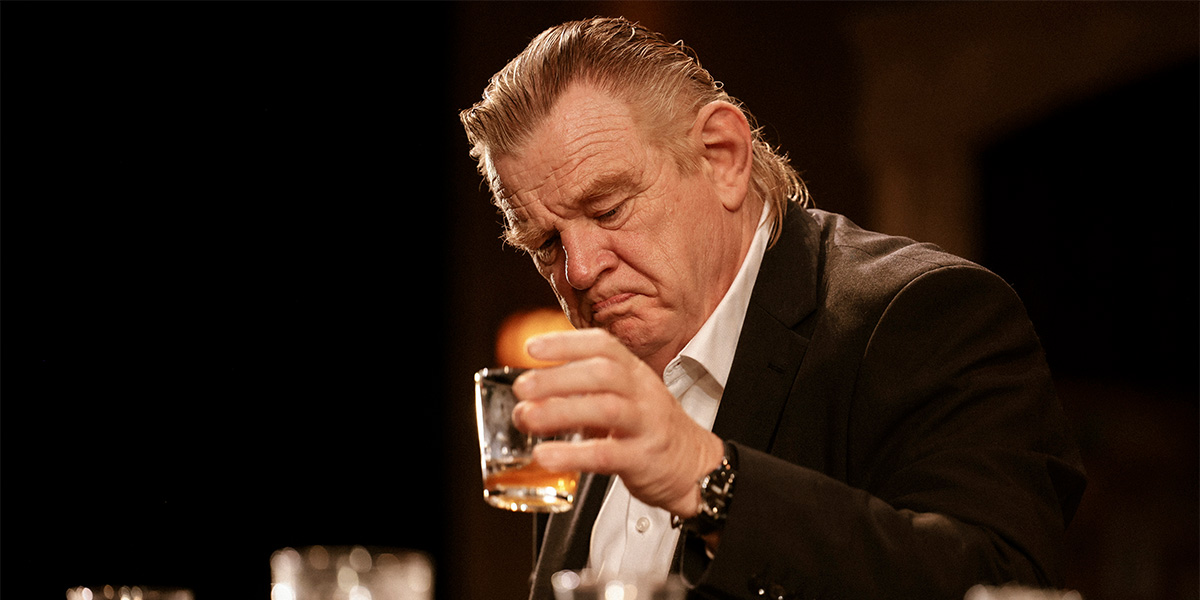Brendan Gleeson as Jack, seated in a dimly lit room, wearing a grey sweater, looking contemplative.