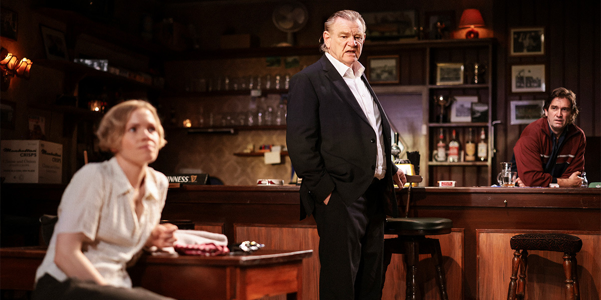Kate Phillips and Brendan Gleeson sit on a wooden bench, engaged in conversation, with a dimly lit pub background.
