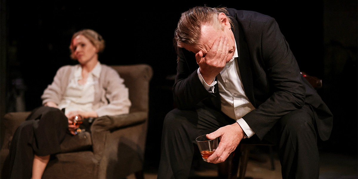 Kate Phillips and Brendan Gleeson sit on a wooden bench, engaged in conversation, with a rustic interior background.
