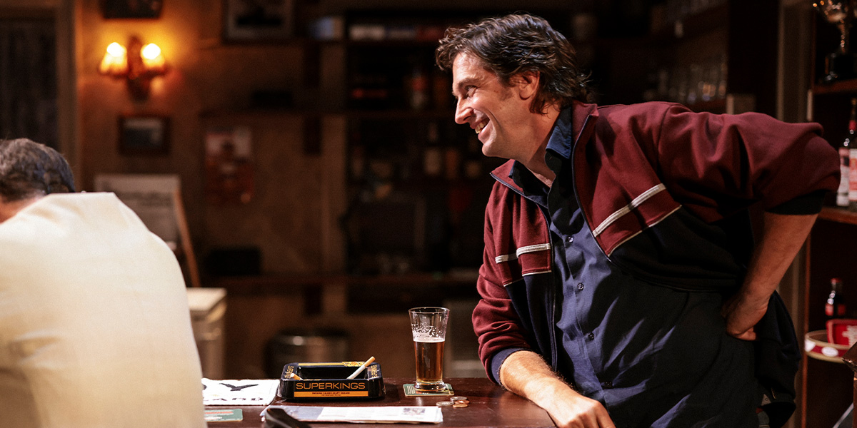 Two men in casual clothing sit at a table, engaged in conversation, with a warm, rustic interior background.