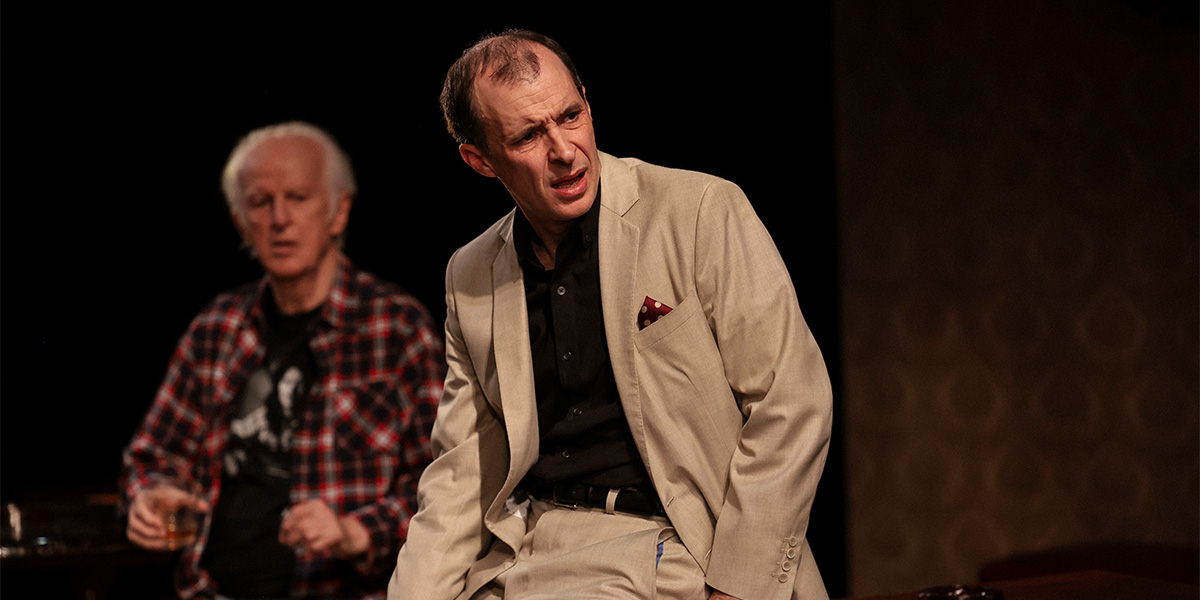 Two men in casual clothing sit at a table, engaged in conversation, with a rustic interior background.