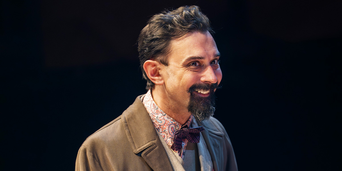 Tom Kanji smiles while wearing a brown coat and patterned shirt, against a dark background.