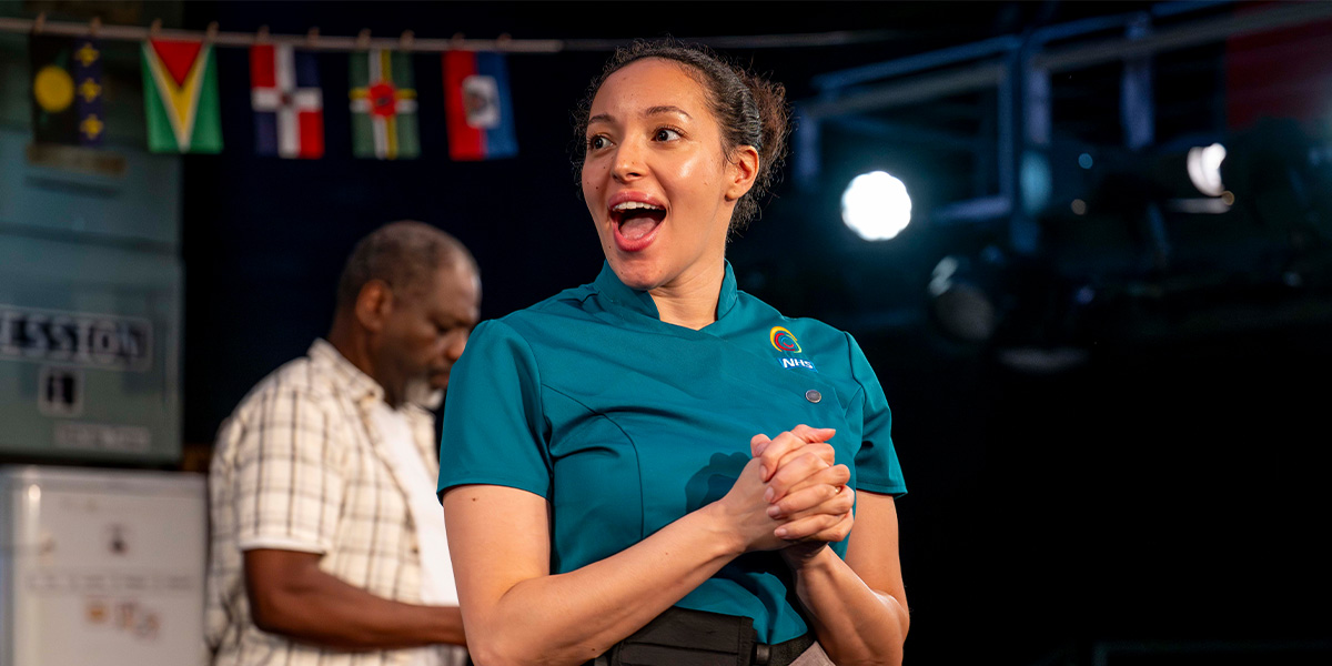Aimee Powell stands in a sunlit room, wearing a patterned dress, smiling and holding a cup.