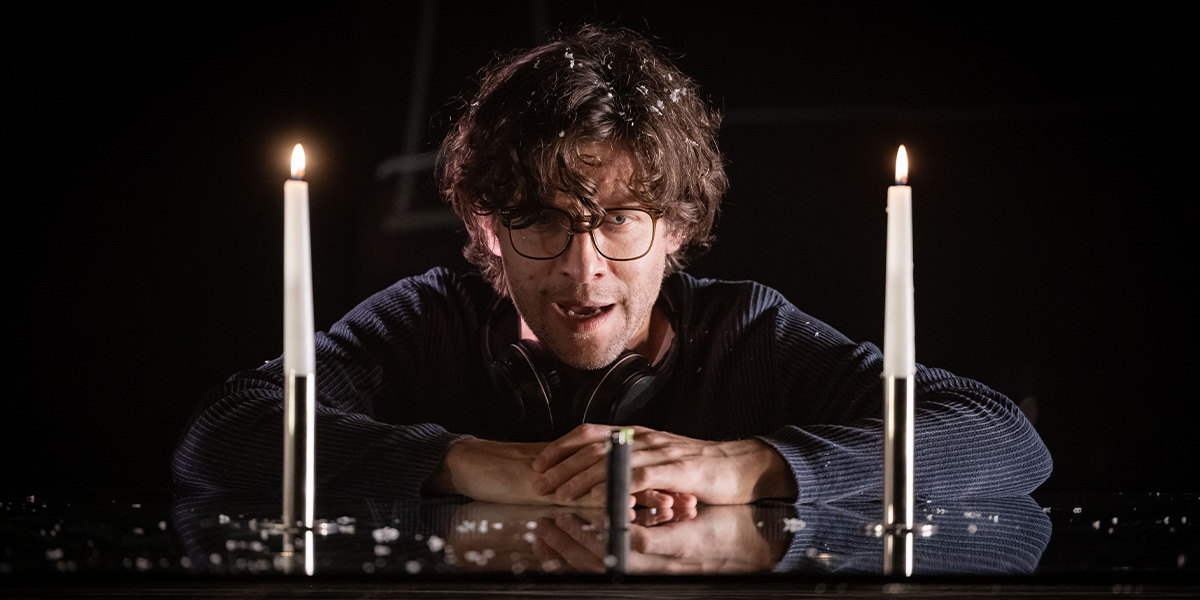 A man with short dark hair, wearing a brown coat, stands in a dramatic pose against a dark background.