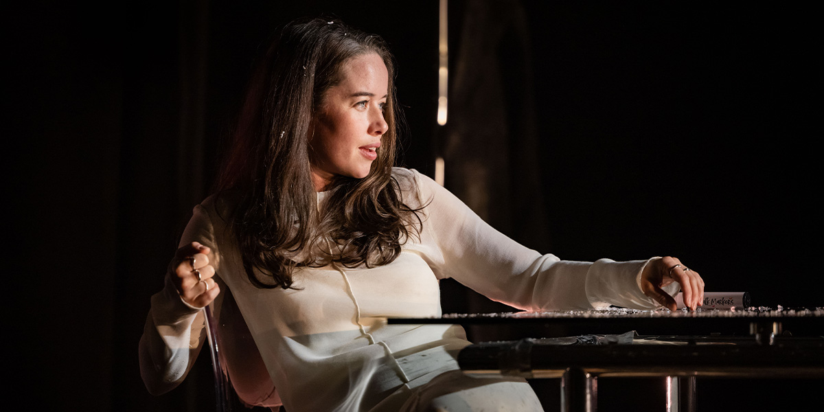 Anna Popplewell as Julia, wearing a dark dress, stands thoughtfully against a blurred outdoor background.