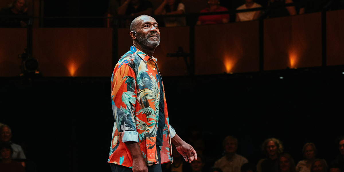 Lenny Henry stands on stage, smiling, wearing a black shirt, with a colourful backdrop behind him.