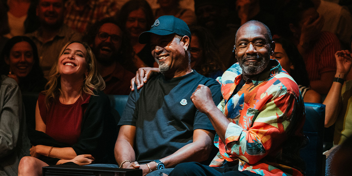 Lenny Henry performing on stage, wearing a black suit, with a colourful backdrop and audience visible.