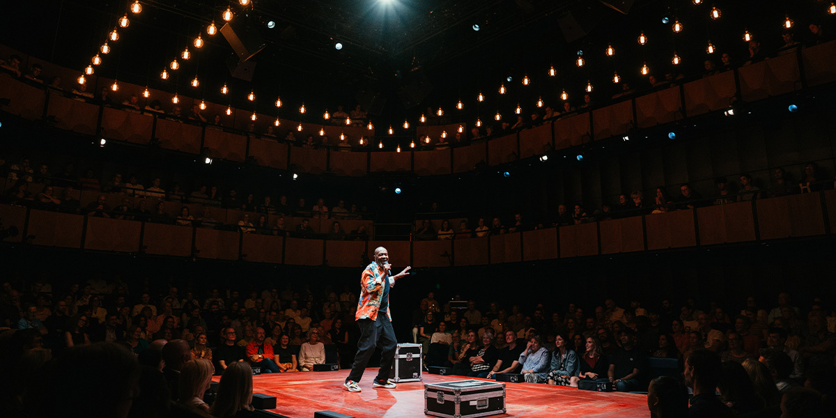 Lenny Henry performs on stage, wearing a black shirt, with a colourful backdrop and audience visible.