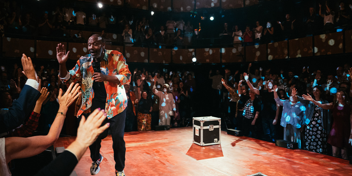 Lenny Henry performs on stage, wearing a blue shirt, with a vibrant backdrop and audience visible.