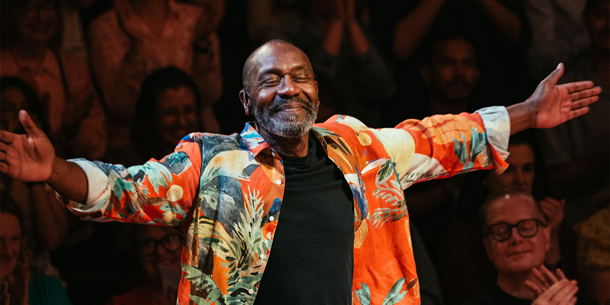 Lenny Henry performs on stage, wearing a blue shirt, with a bright spotlight and audience in darkness.