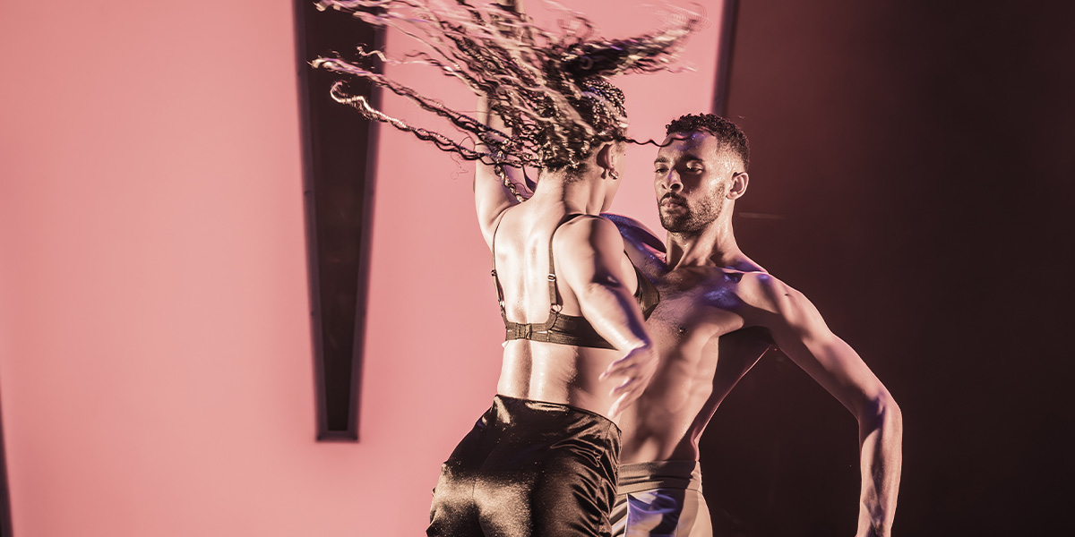 Two performers in dynamic movement under pink lighting, one with long braided hair swinging dramatically.