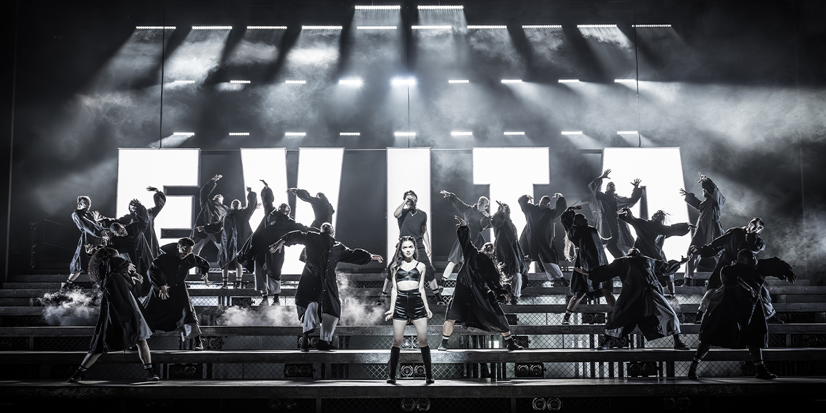 Dynamic stage performance featuring multiple people in black outfits, centred around a prominent singer, beneath bright overhead lights.