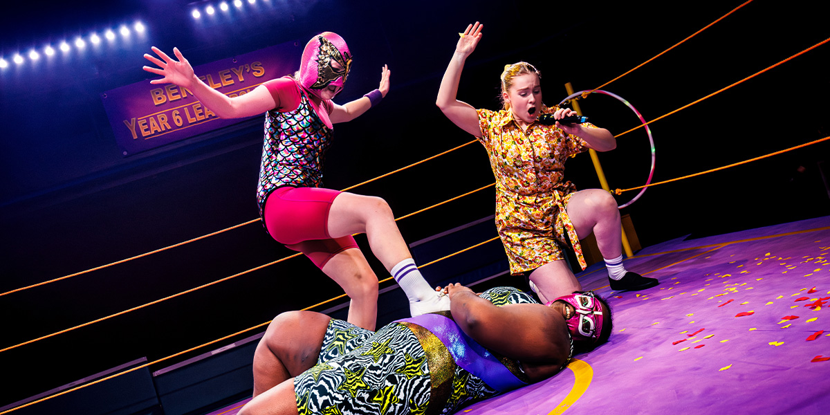 A group of four people in colourful costumes poses enthusiastically at a promotional event, with banners in the background.