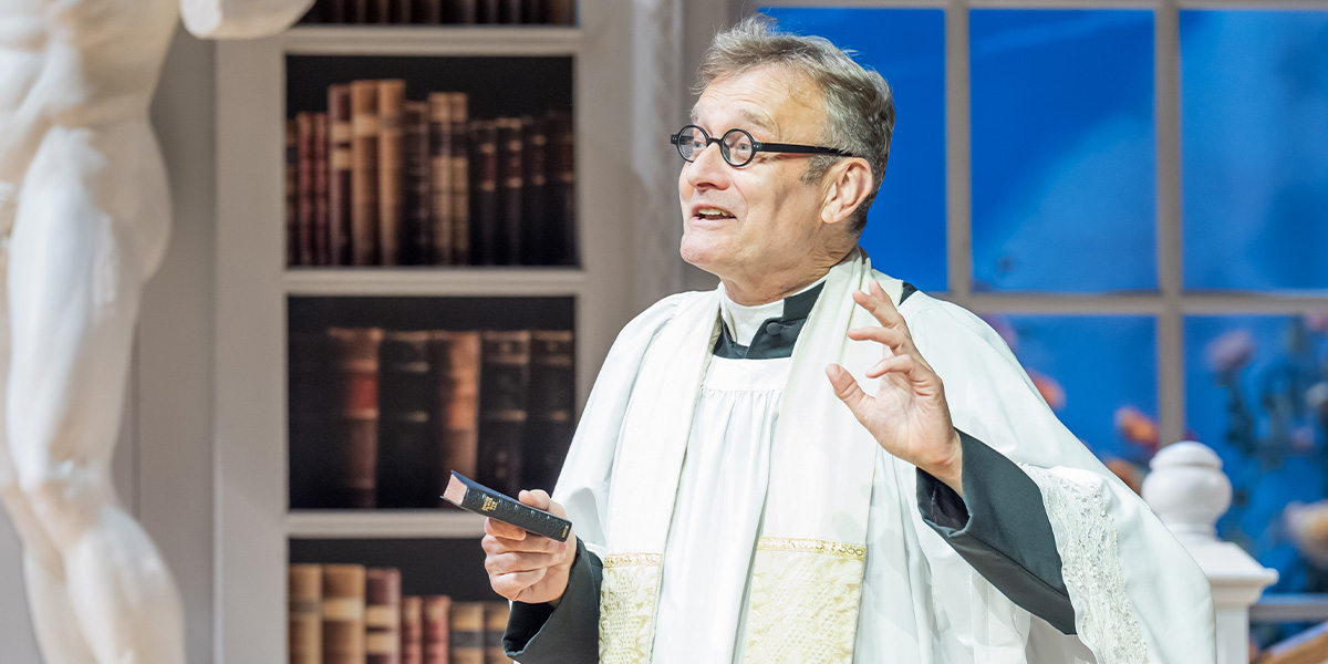 Hugh Dennis, dressed in a formal suit, smiles while holding a script on a theatre stage.