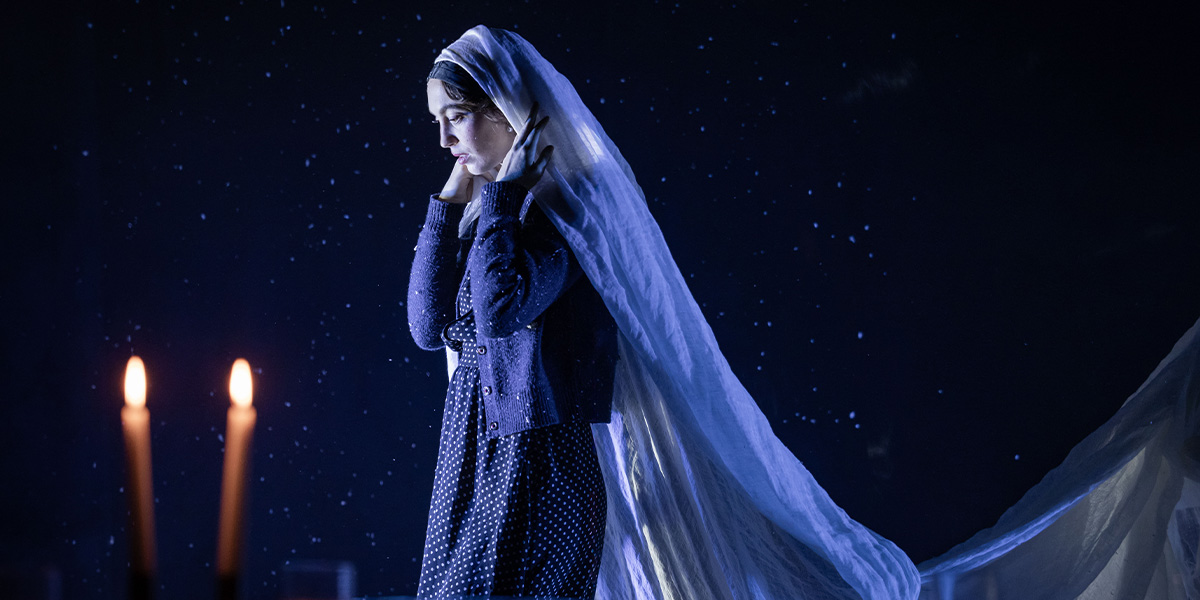 Katerina Tannenbaum as Esther, wearing a flowing blue dress, stands confidently against a dark, textured backdrop.