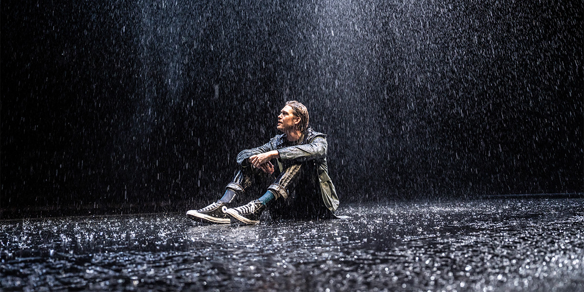 A man with light brown hair, wearing a dark suit, stands on a rocky shore, gazing thoughtfully at the sea.