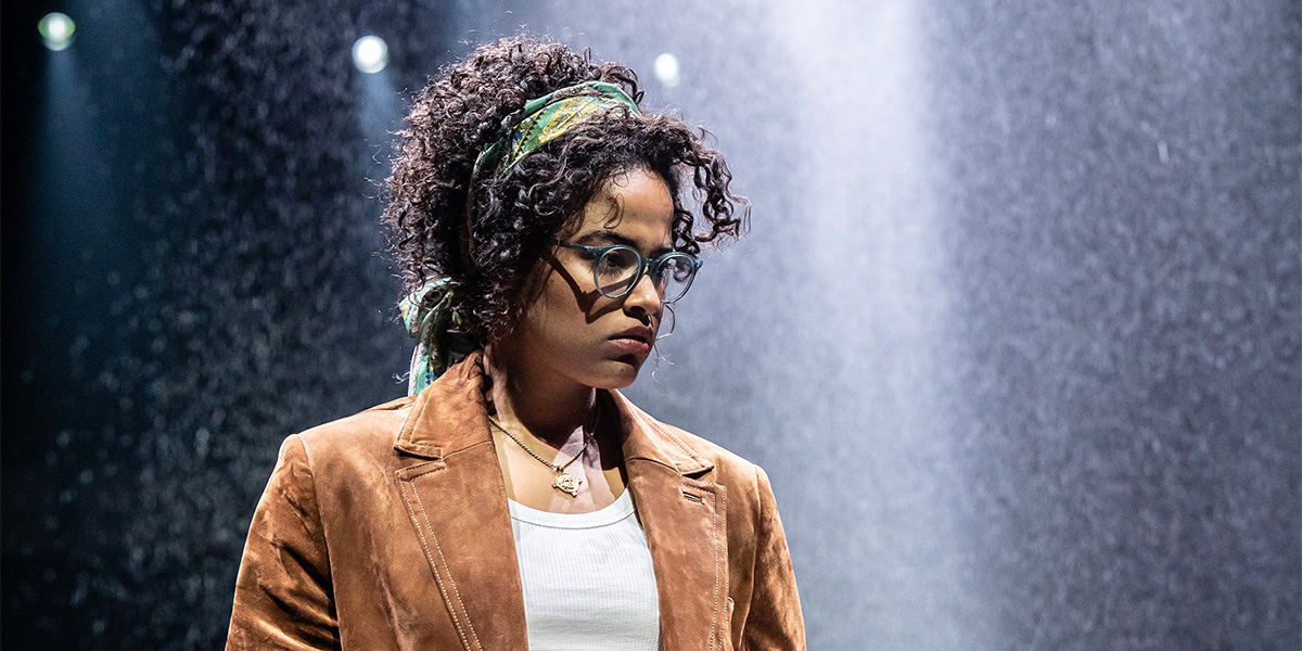 Two actors in period costumes, one in a flowing dress, the other in a suit, on stage.