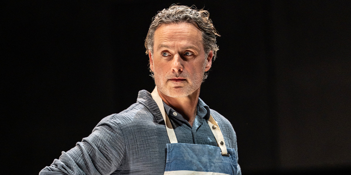 A man with dark hair and a beard stands in a dimly lit room, looking contemplative.