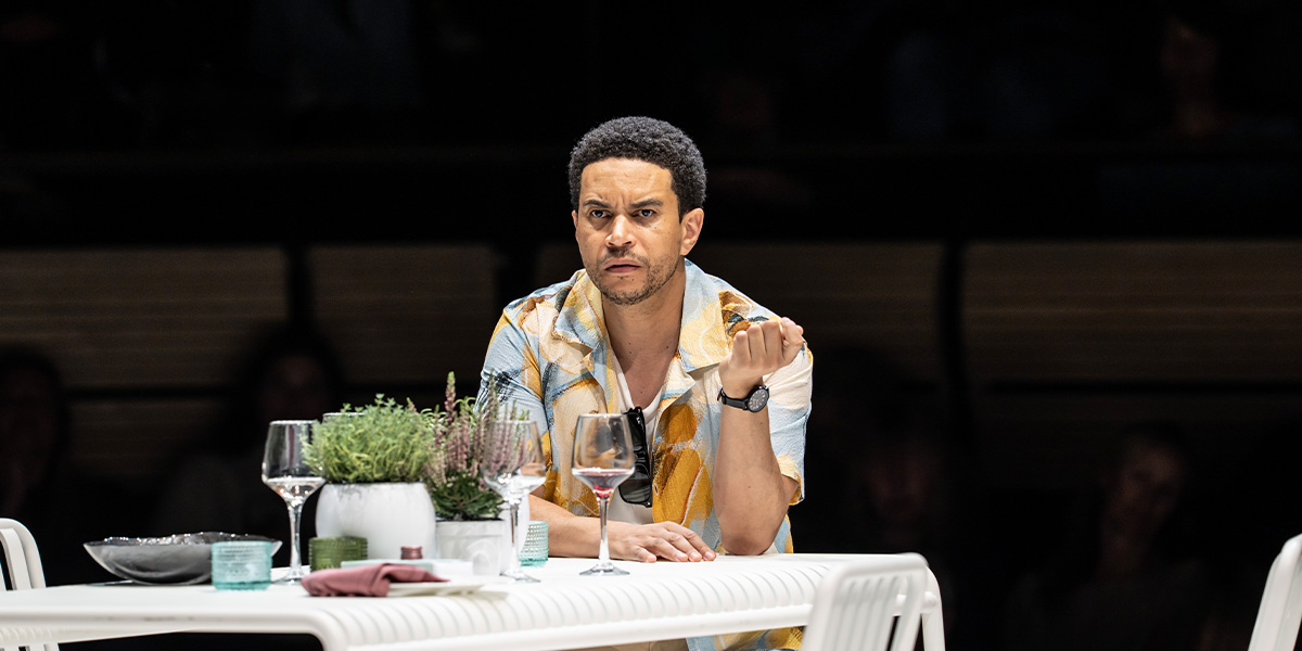 A man in a dark suit stands on a rocky shore, gazing thoughtfully at the sea.