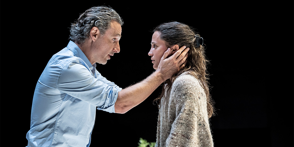 Andrew Lincoln and Alicia Vikander are seated together, smiling, against a softly lit, neutral background.