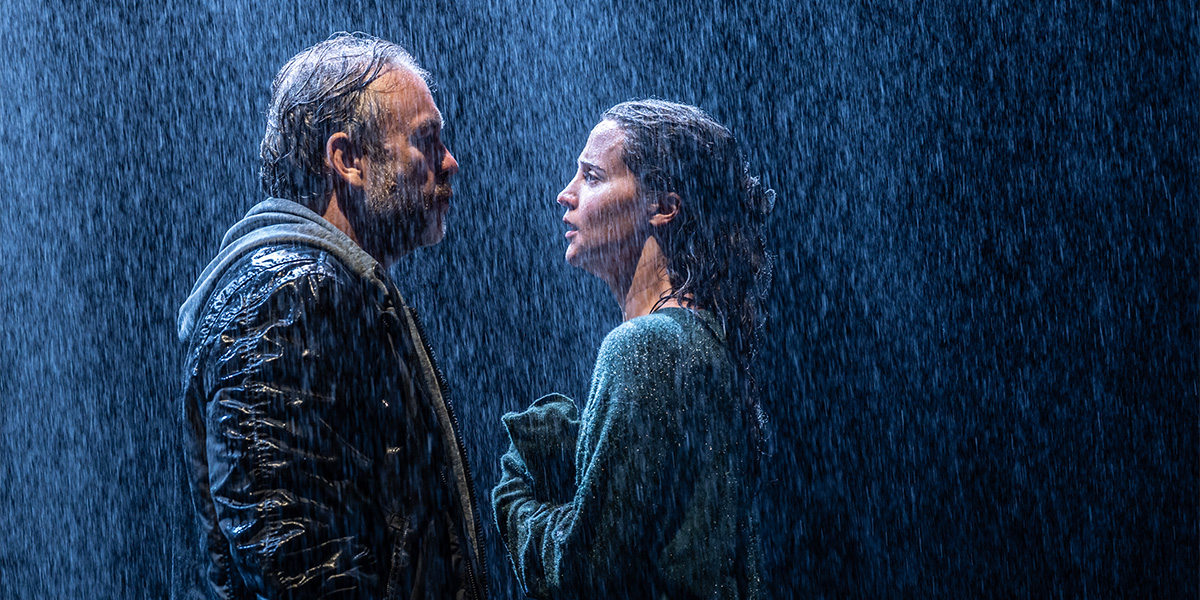 Brendan Cowell and Alicia Vikander pose together, smiling, against a backdrop of soft blue ocean waves.