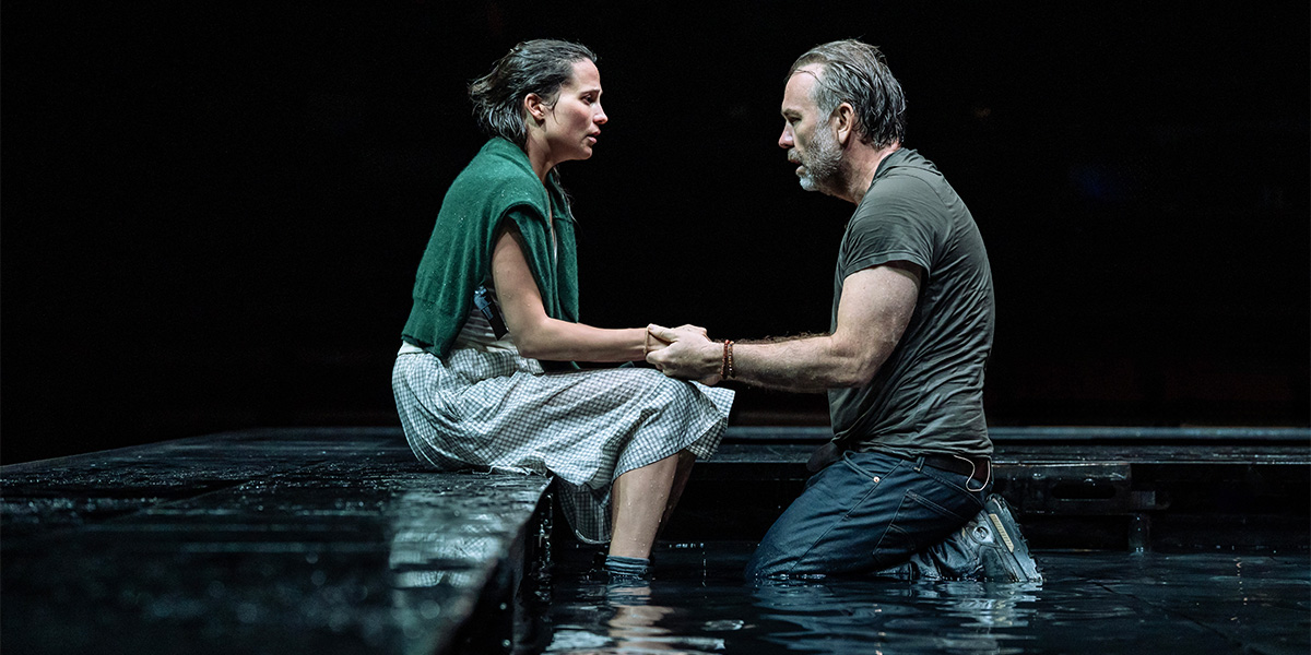 Alicia Vikander and Brendan Cowell sit together, engaged in conversation, surrounded by a coastal backdrop.