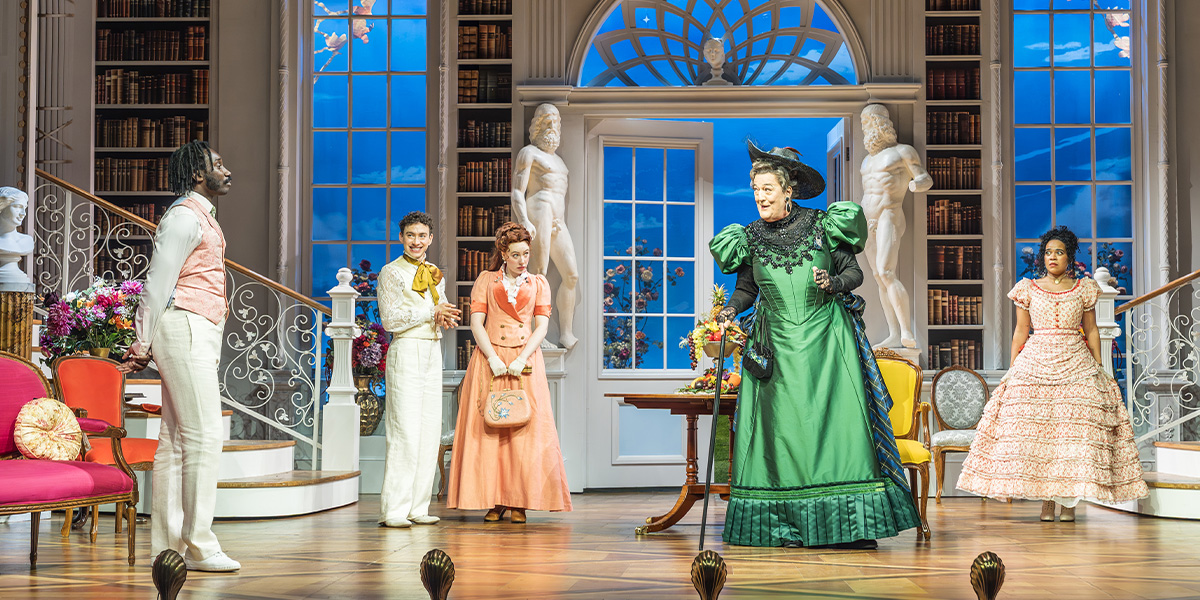 Five actors in formal attire pose together, smiling, with a theatre backdrop featuring ornate decorations.