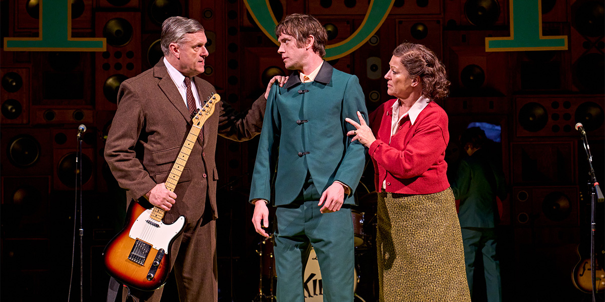 Three men in casual clothing perform on stage, with warm lighting and a vibrant backdrop.